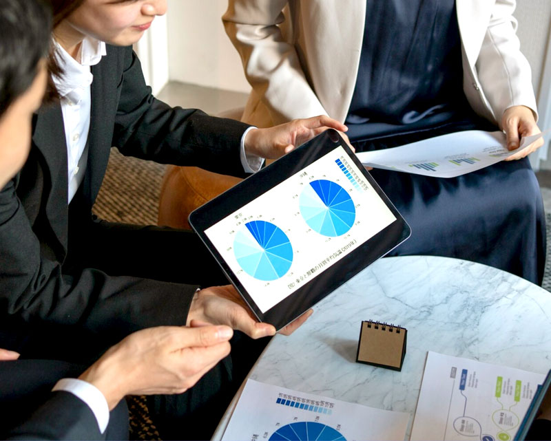 Three people in business attire are sitting around a table reviewing charts and graphs. One person is holding a tablet displaying pie charts, while another person is holding a document with bar charts.